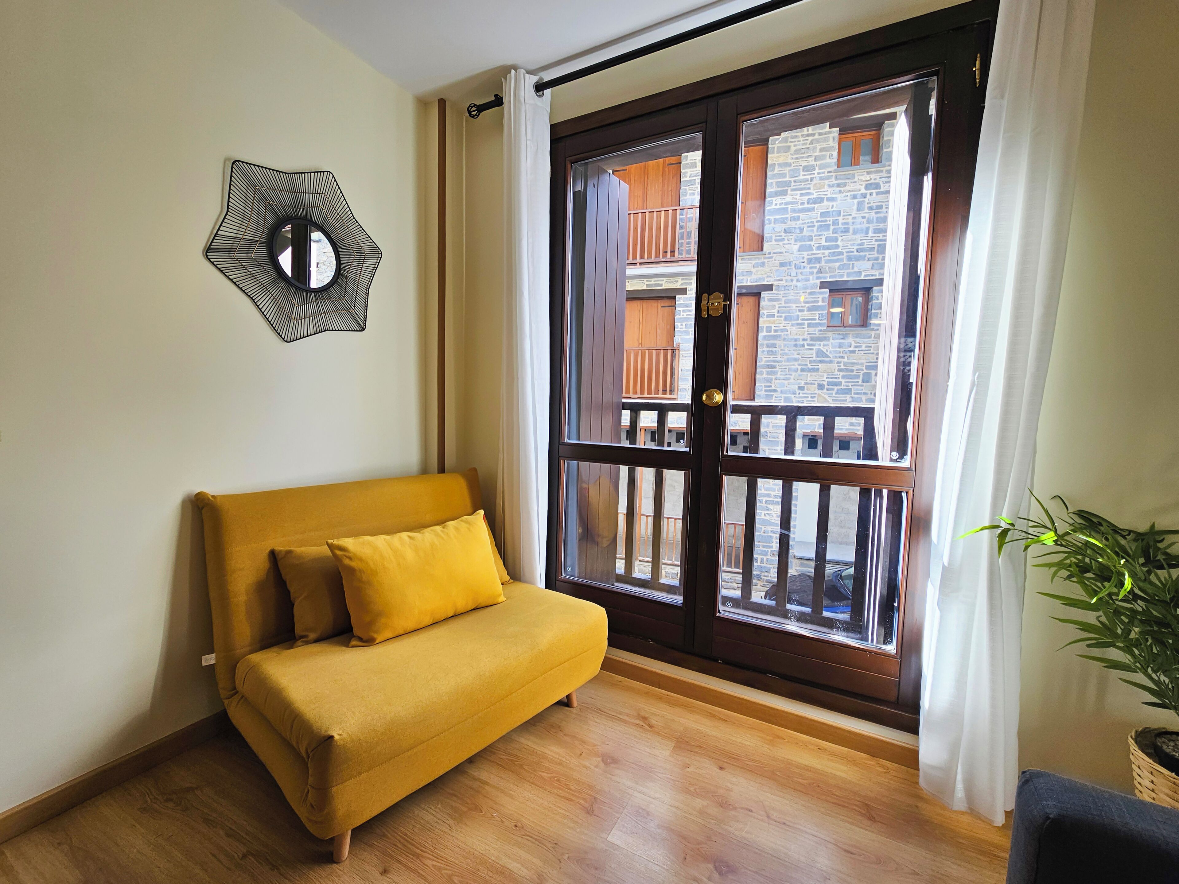 Une pièce avec un canapé jaune, un plancher en bois, une grande fenêtre donnant sur un balcon et un élément décoratif fixé au mur.