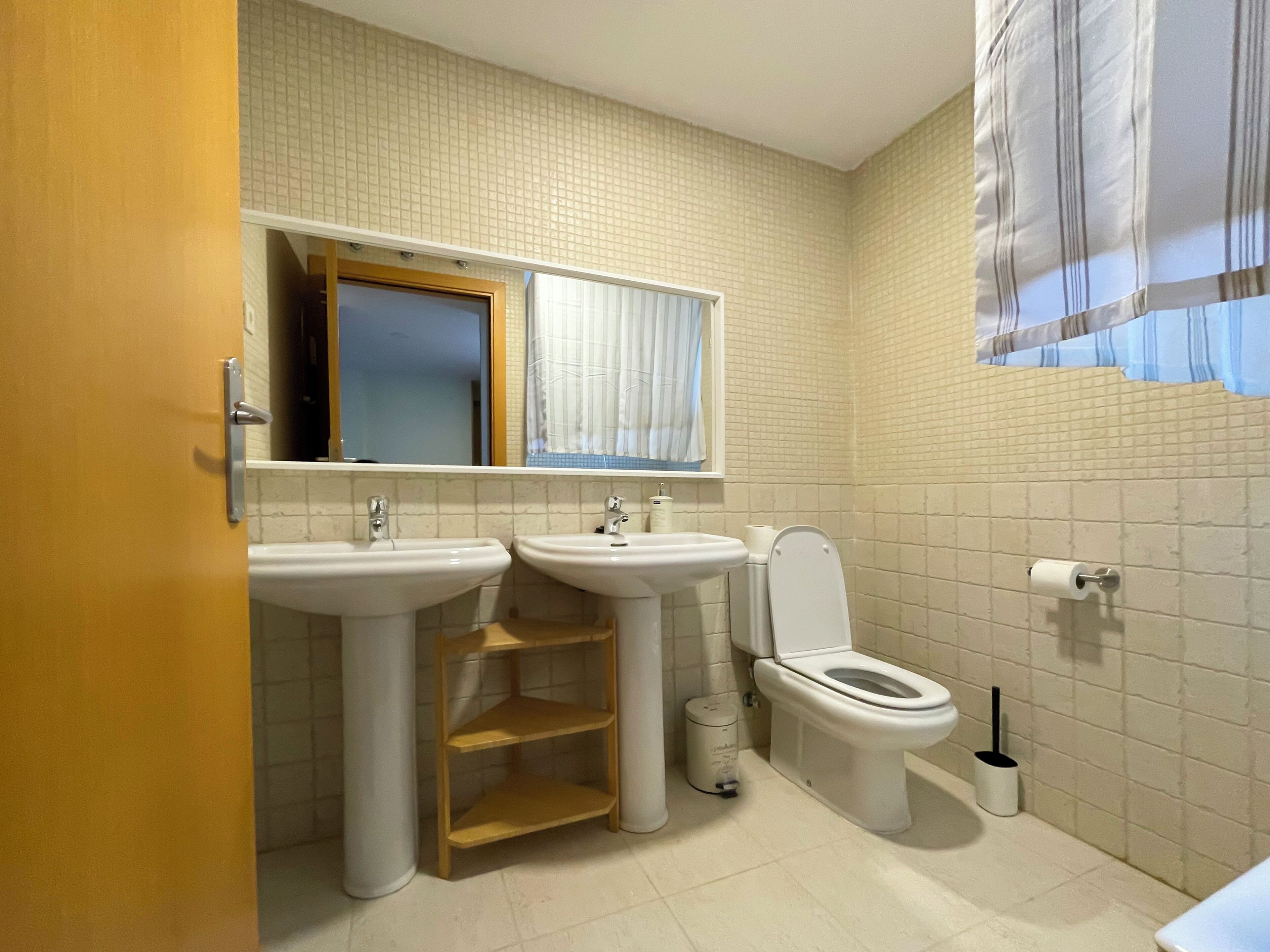 Un baño con dos lavamanos, un inodoro, un espejo y una ventana con cortinas.
