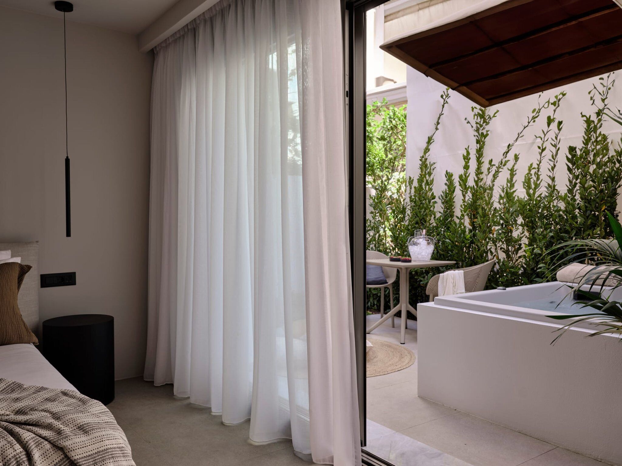 A modern bedroom with a large window, white curtains, a bed, a bedside table, and a view of an outdoor patio with plants and a table.