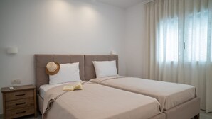 Una cama doble con cabecera color beige y dos almohadas, una mesita de noche con lámpara, y una ventana con cortinas translúcidas.