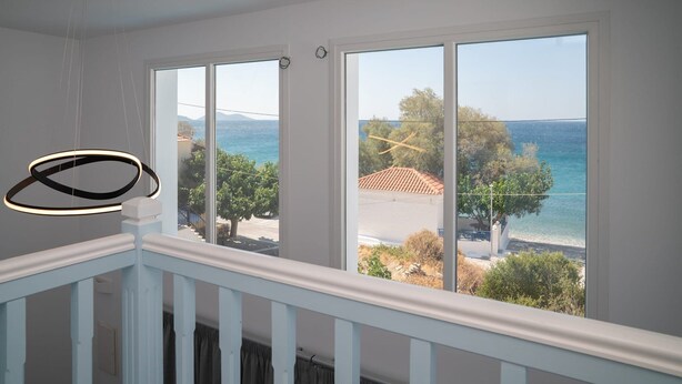 Habitación con amplias ventanas que dan a una playa y al mar, barandilla blanca y lámpara moderna.