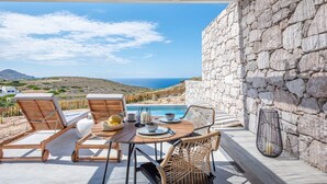 A terrace with a stone wall, wooden lounge chairs, a table set for two, and a view of the sea and hills. - Dazzling Milos Suite | Elysian Suite | Panoramic Views of Sea and Milos (Milos)