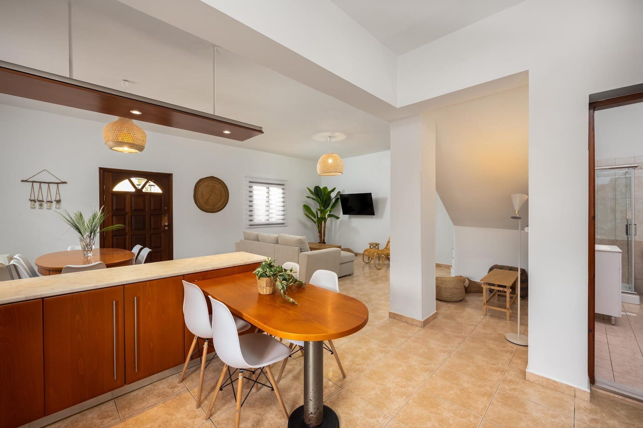 A modern kitchen with a wooden island, white chairs, and a dining table. There is a TV mounted on the wall, a sofa, and a plant in the living area.