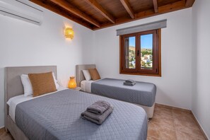 A bedroom with two beds, a window with a view, and a wall-mounted air conditioner. - Dazzling Symi Villa | Serenity House | 3 Bedrooms | Breathtaking City Views |. (Symi)