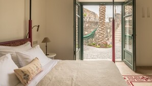 Une chambre à coucher avec un lit, une table de chevet, une lampe et une vue sur un mur de pierre et un hamac à l’extérieur.