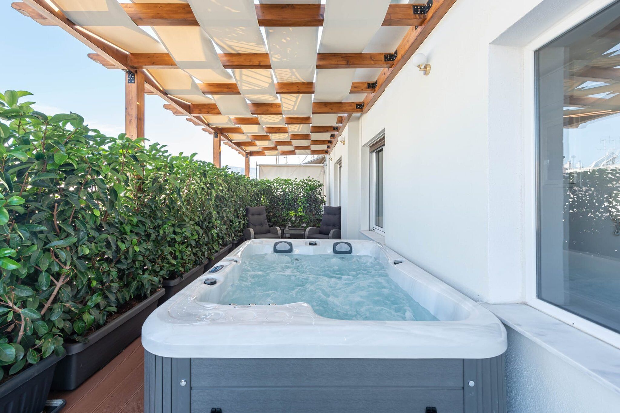 A hot tub under a wooden pergola, surrounded by lush greenery and a clear view of the sky.