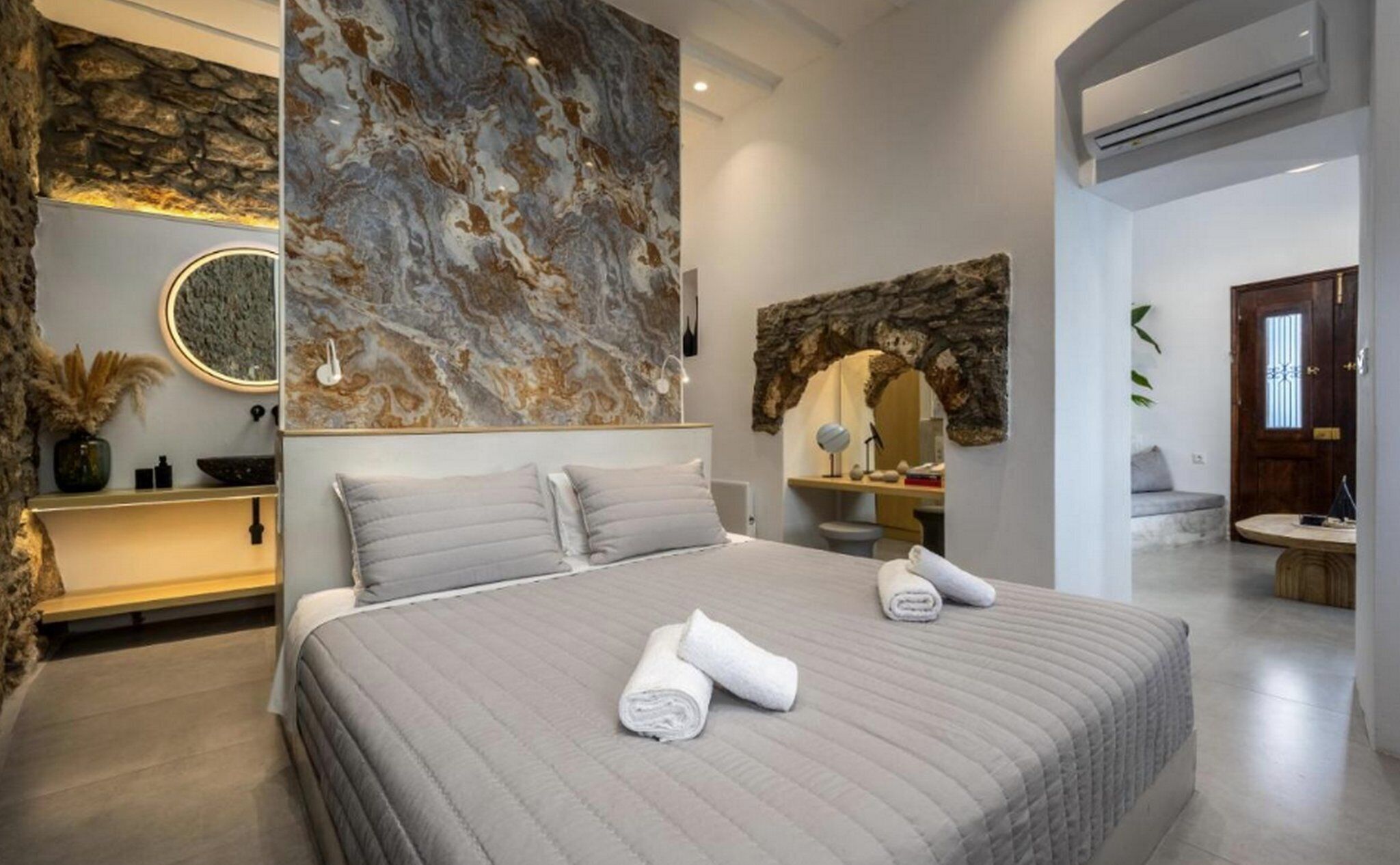 Ein modernes Schlafzimmer mit einem großen Bett, einer Marmorwand, einer Wand mit Steinakzenten, einem runden Spiegel und einem kleinen Schreibtisch.