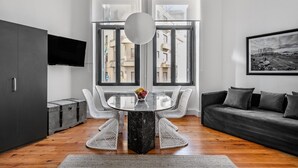 Un salon moderne avec une table ronde en verre, des fauteuils en osier blancs, un canapé foncé et un téléviseur fixé au mur.