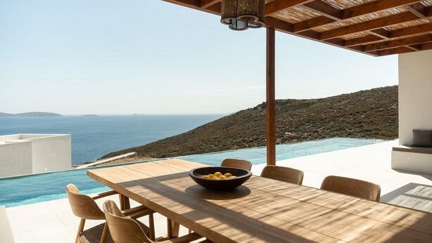 Een houten tafel met stoelen en een fruitschaal onder een pergola, met uitzicht op zee en een bergachtig landschap.