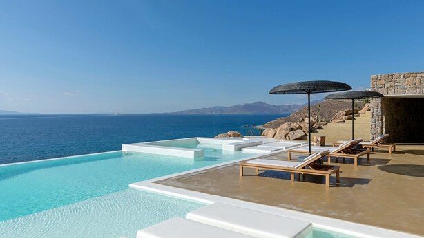 Une piscine à débordement à l’eau d’un bleu cristallin, avec des chaises longues blanches et des parasols, offrant une vue imprenable sur une côte montagneuse.