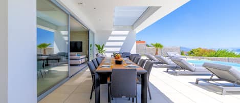 A modern outdoor dining area with a long table, chairs, and a poolside lounge with sun loungers.