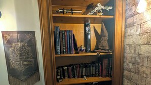 A wooden bookshelf with books and decorative items, a framed poster on the wall, and a stone-textured wall. - The Sorcerer's Den (Caumont-sur-Durance)