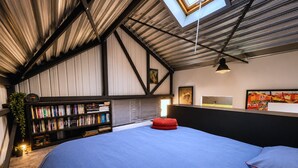 A bedroom with a large bed, a bookshelf, a red pouf, and a skylight.