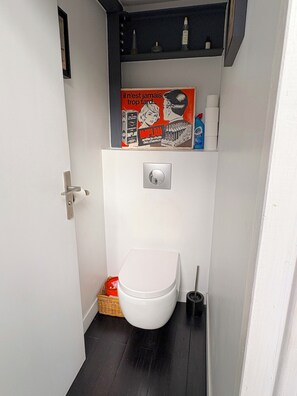 Ein kleines Badezimmer mit einer weißen Toilette, einem Regal mit Pflegeprodukten und einer Zeitschrift sowie einem kleinen Korb am Boden.