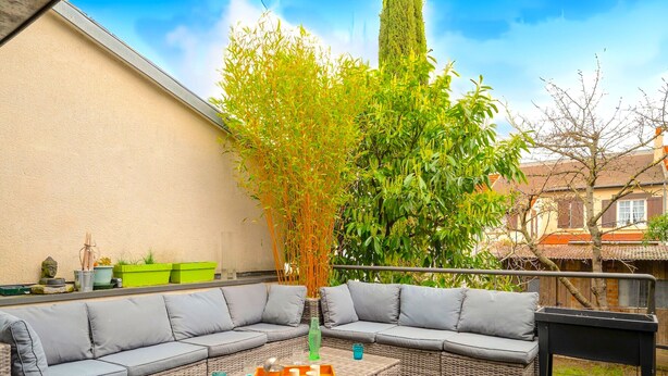 An outdoor seating area with wicker sofas and a coffee table, surrounded by greenery and a wooden deck.