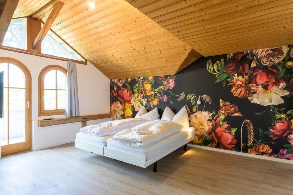 Une chambre avec un plafond en bois, un grand lit, une fresque murale à motifs floraux et une fenêtre avec des rideaux.