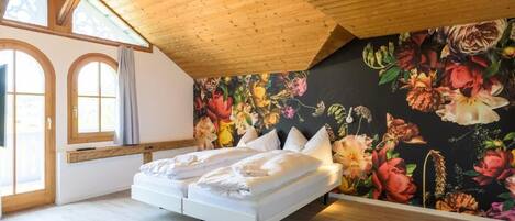 A bedroom with a wooden ceiling, a large bed, a floral mural, and a window with curtains.