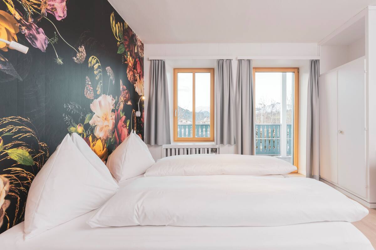 Ein Schlafzimmer mit Bett, Heizkörper, einem Fenster mit Vorhängen und einem Balkon mit Geländer.