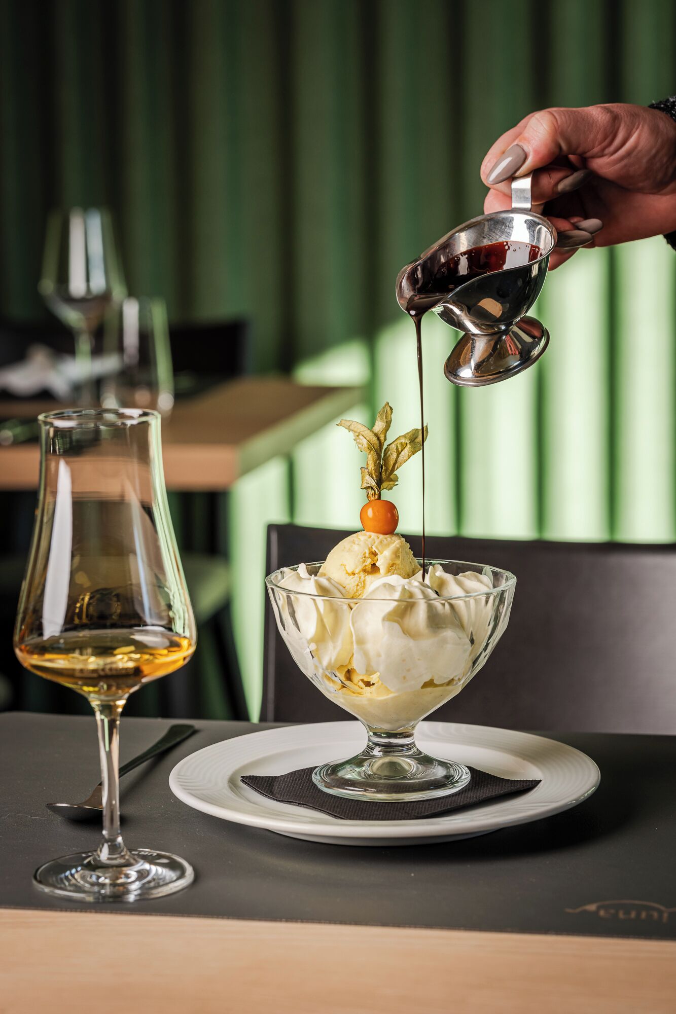 Un dessert composé de crème glacée et d’une cerise, le tout présenté sur une assiette, un verre de vin, et une main versant du sirop.