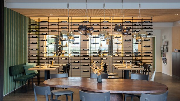 A modern dining area with a wooden table, grey chairs, and a wine rack in the background.