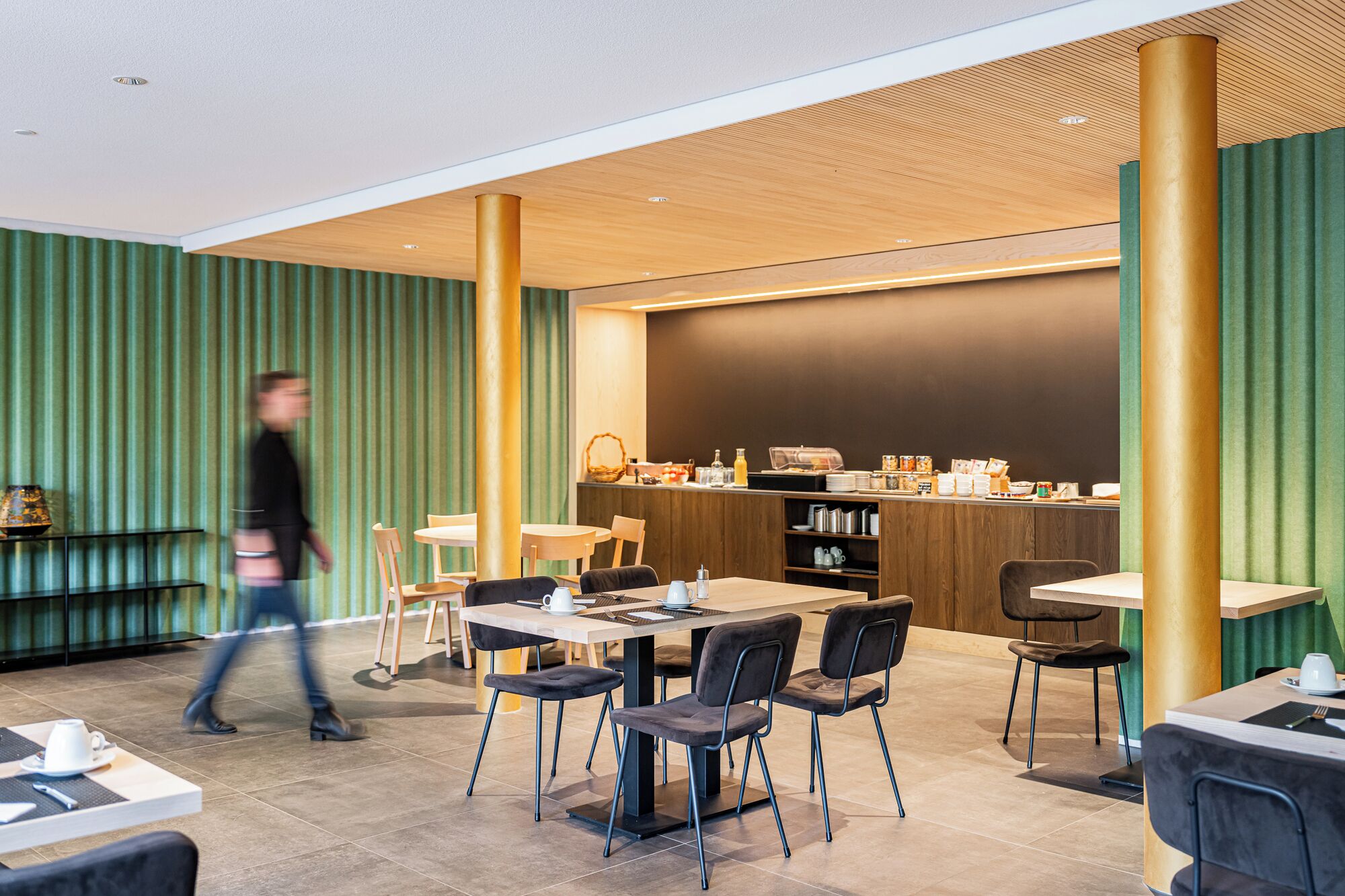 Un café moderno con techo de madera, paredes verdes y una barra con productos para el desayuno.
