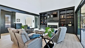 Ein modernes Wohnzimmer mit Sofa, Sesseln, Couchtisch und Kamin. Regale mit Dekorationselementen und ein großes Fenster mit Blick.