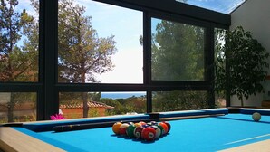 A pool table with a colorful arrangement of balls, large windows with a view of trees and a building, and a potted plant to the right. - L'ESCALE COTE BLEUE VILLA & Spa 4étoiles (Sausset-les-Pins)