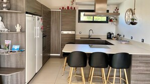 A modern kitchen with a central island, bar stools, a built-in oven, and a window above the sink. - L'ESCALE COTE BLEUE VILLA & Spa 4étoiles (Sausset-les-Pins)