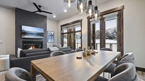 Una sala de estar moderna con chimenea, sofás grises, una mesa de comedor de madera y ventanas amplias que ofrecen vistas a un paisaje nevado.