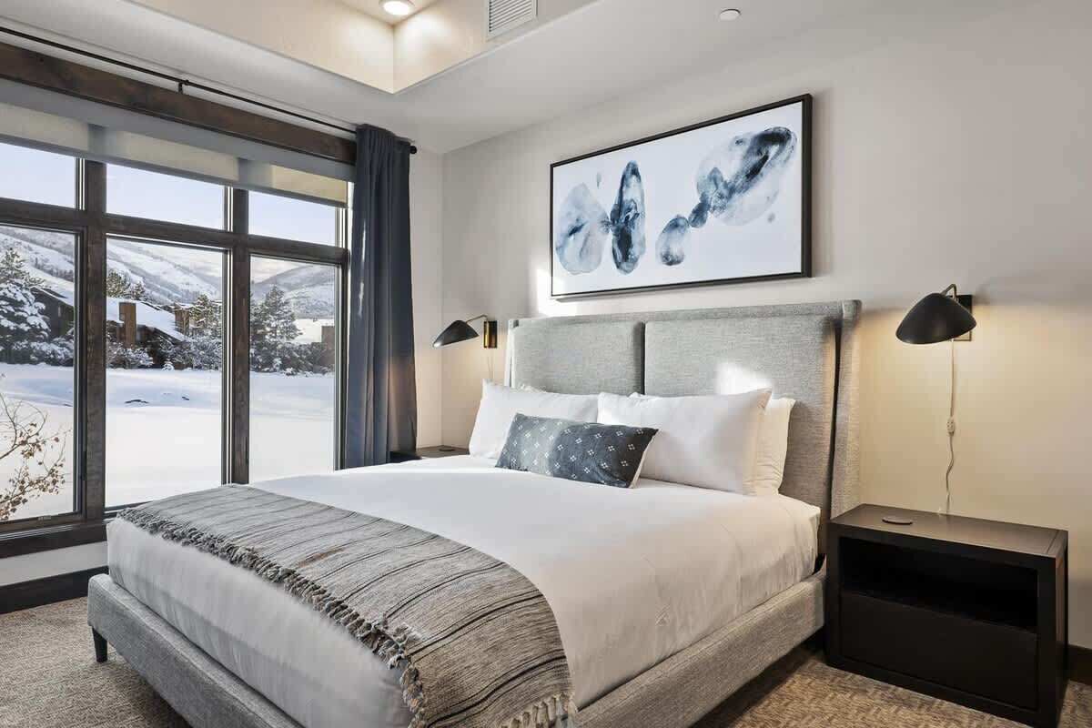 A modern hotel room with a large bed, a view of a snowy landscape, and a framed abstract painting on the wall.