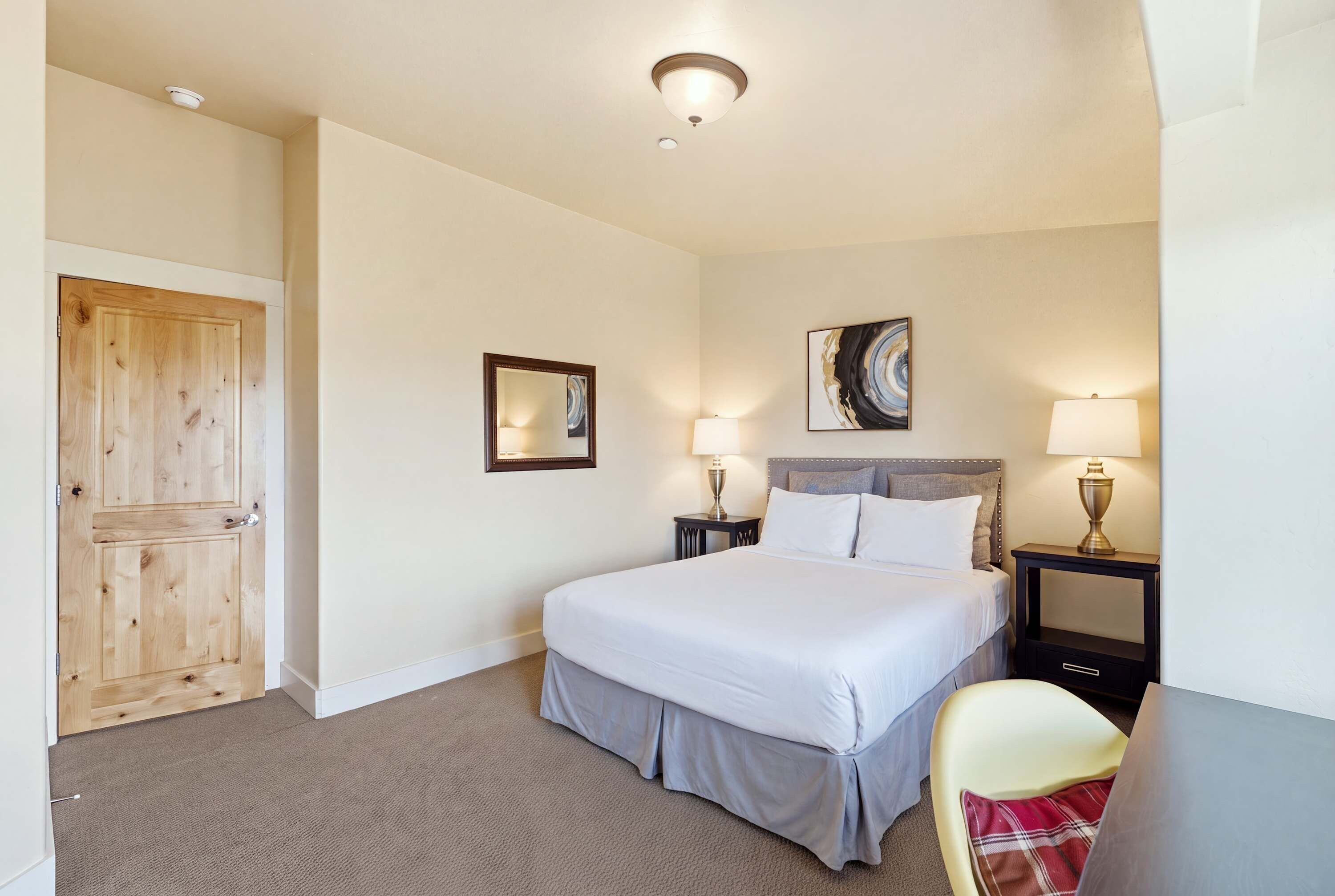 A hotel room with a bed, two bedside tables with lamps, a wooden door, a framed picture on the wall, and a carpeted floor.