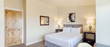 A hotel room with a bed, two bedside tables with lamps, a wooden door, a framed picture on the wall, and a carpeted floor.