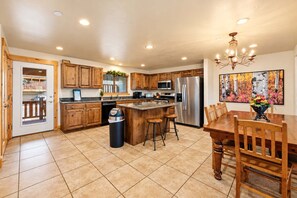 A modern kitchen with wooden cabinets, stainless steel appliances, a central island with stools, and a dining area with a chandelier. - Bears Lair | Private Hot Tub & Mountain Views (Park City)