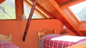 Un dormitorio de cabaña de madera con techo inclinado, una cama con colcha escocesa, una mesita de noche con lámpara y una ventana con vistas a una montaña boscosa.