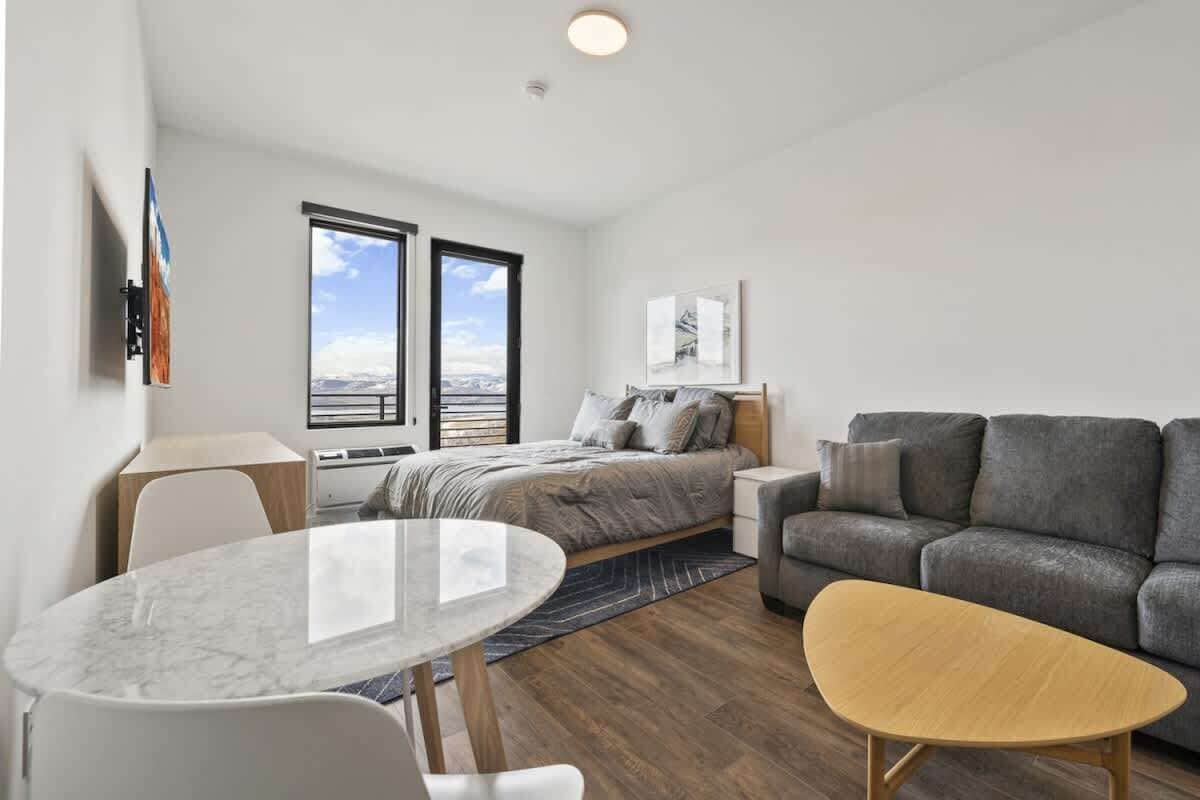 Un dormitorio moderno con cama, un sofá gris, una mesa redonda y un ventanal con vista a la ciudad.