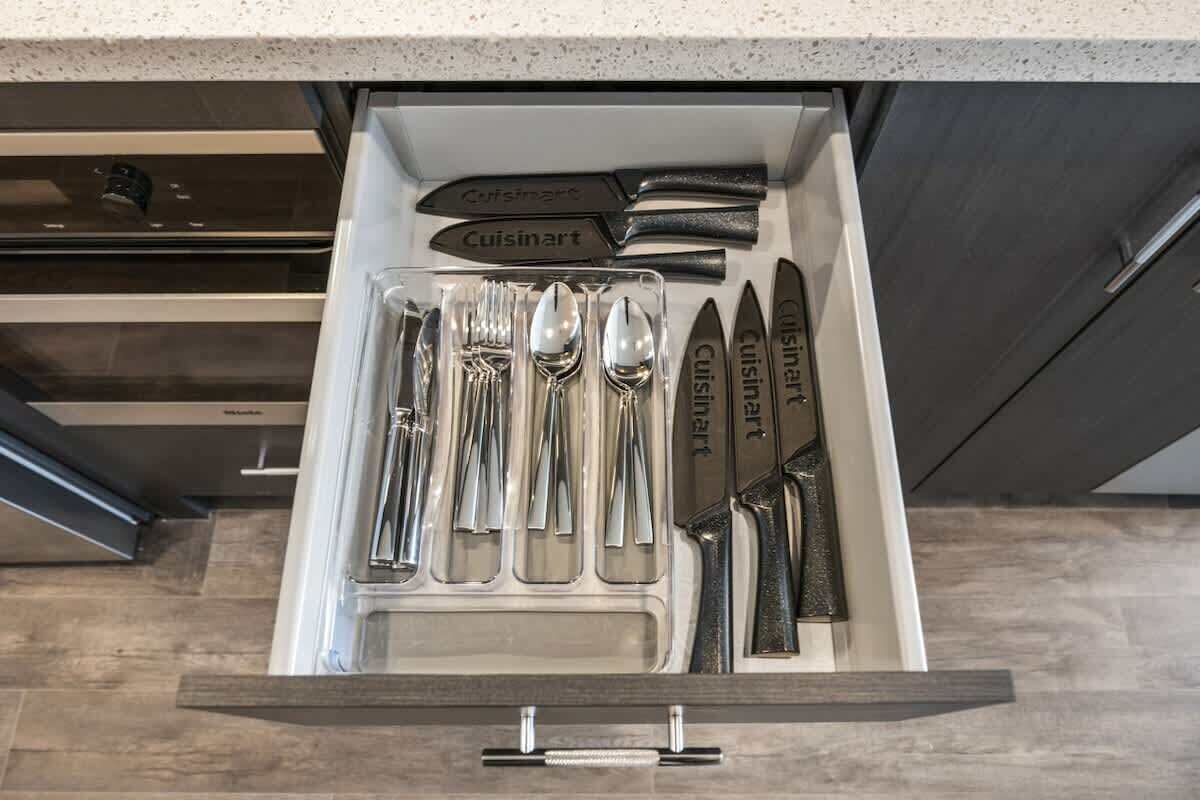 A drawer containing a set of silverware labeled 'Cuisinart' with a modern kitchen backsplash and wooden flooring.