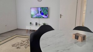 Un salon moderne avec une table à manger en marbre, des chaises noires, un téléviseur à écran plat fixé au mur, une petite table d’appoint blanche et une horloge.