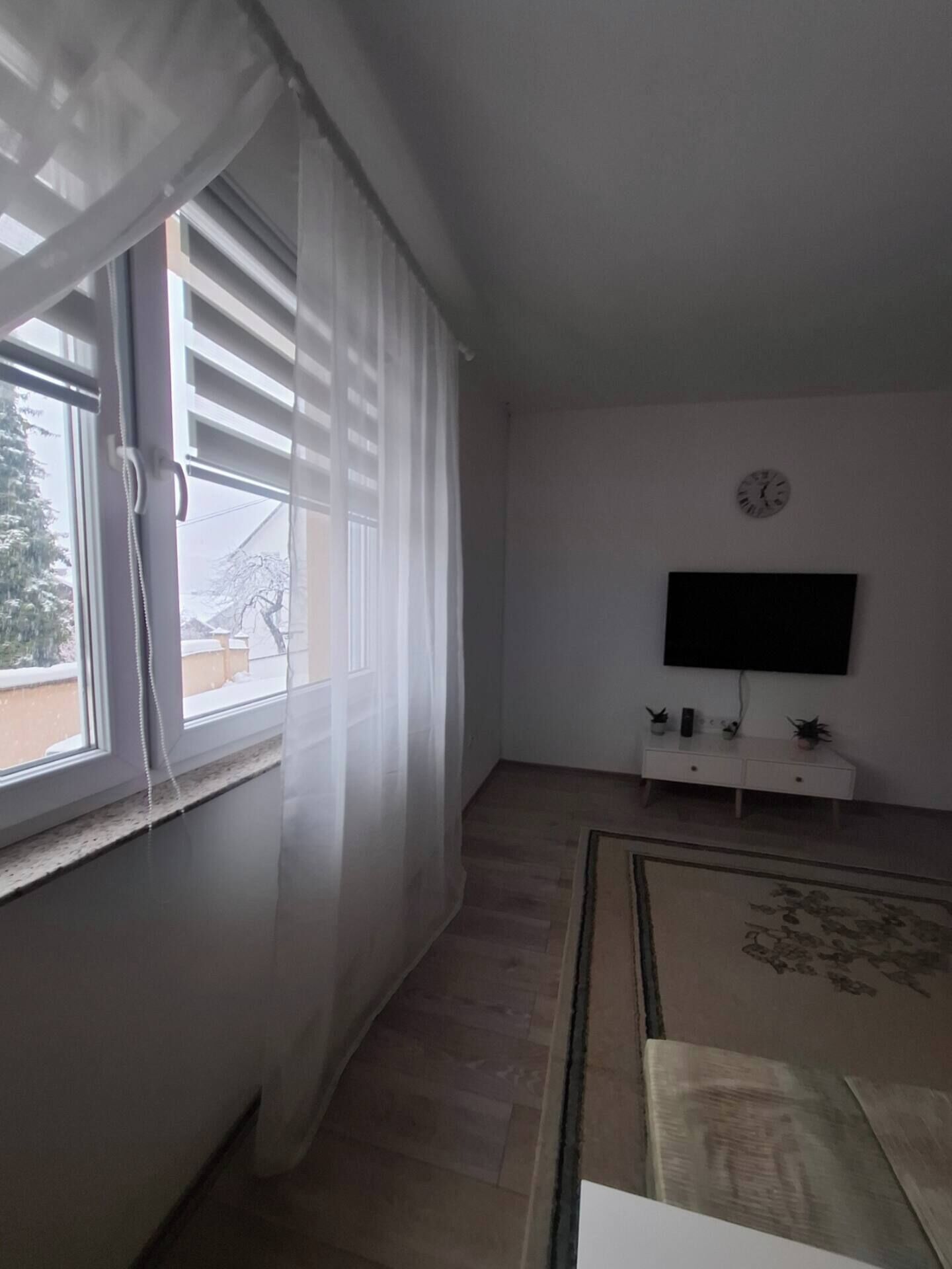 Ein Zimmer mit großem Fenster, bedeckt mit transparenten Vorhängen, einem wandmontierten Flachbildfernseher, einem kleinen weißen Schrank darunter und einem gemusterten Teppich auf dem Boden.