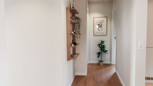 Un couloir avec une étagère en bois adossée au mur, une œuvre d’art encadrée au-dessus, et une plante en pot, visible au loin.