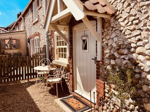 A stone house with a white door, a small porch with a table and chairs, and a wooden fence. - Sutton Cottage - sleeps 2 guests  in 1 bedroom (Burnham Market)