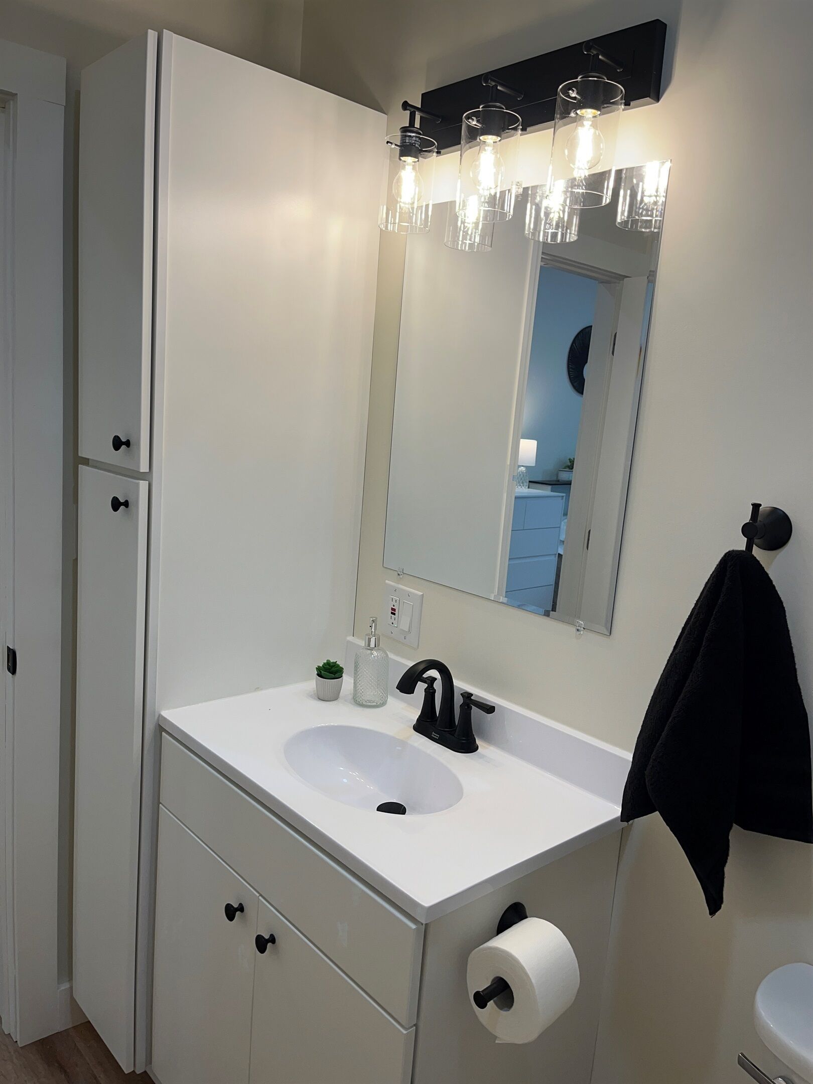 A modern bathroom with a white sink, black faucet, and a mirror above it. There is a towel hanging on the wall and a small potted plant on the counter.