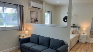A modern living room with a dark grey sofa, a white bedside table with a lamp, a bed with a white duvet, and a large window with curtains. - Full Renovation, new construction throughout. 8 of 8 Studios, 1 mile to ASV (Oneonta)