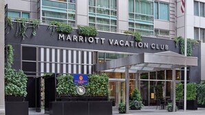 The exterior of the Marriott Vacation Club building with a modern design, featuring a large entrance with glass doors, a covered outdoor seating area, and greenery on the facade.