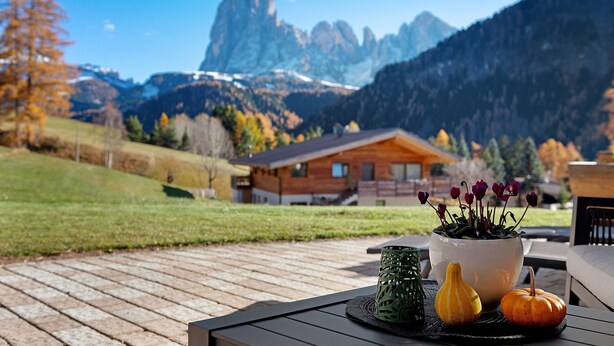 Eine Holzterrasse mit Tisch, einer Topfpflanze und Blick auf Berge sowie eine Holzhütte.