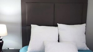 A bed with a dark headboard and white pillows, a blue bedspread, and a bedside table with a lamp.
