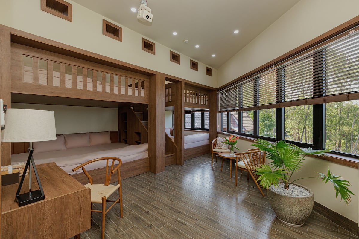 Une chambre avec un lit superposé, un bureau avec une lampe, une chaise, une plante en pot et une fenêtre avec des stores.