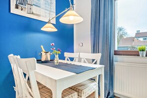 A dining area with a white table, chairs, a blue wall, and a window with a view of houses. - Apartment '1' with Modern Country-Style Kitchen (Cuxhaven)