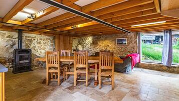 Un intérieur rustique avec un mur en pierre, des poutres en bois au plafond, une cheminée et un coin repas comprenant des chaises et une table.