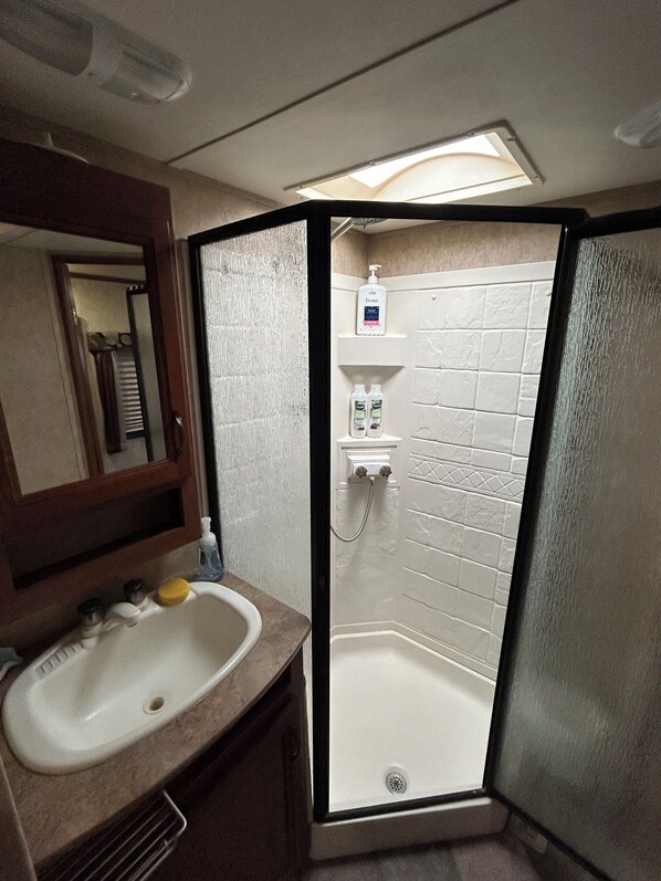 A bathroom with a sink, mirror, toiletries, and a shower with a frosted glass partition. - Blue Lagoon. Enchanting RV in sunny Naples (Naples)