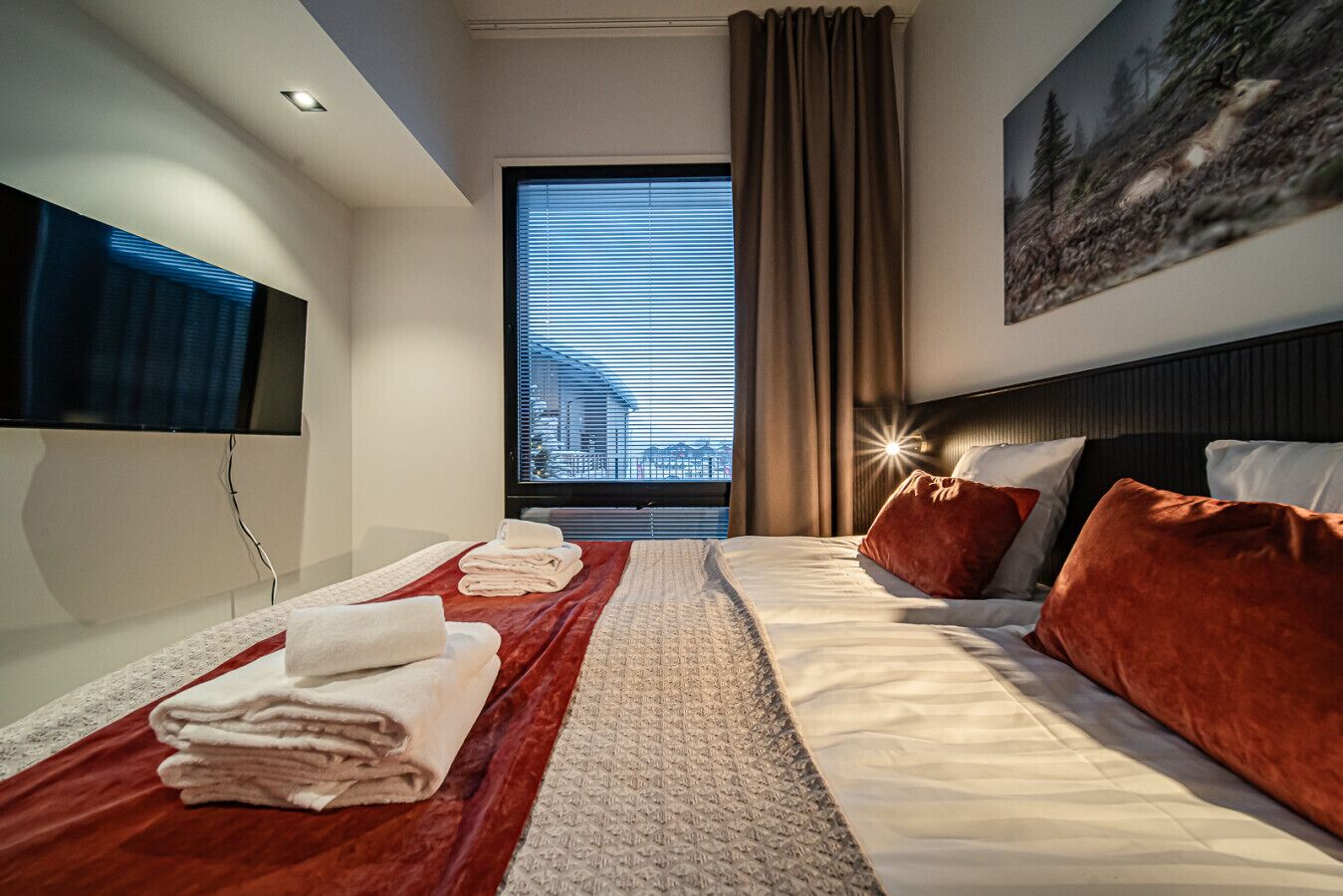 A modern hotel room with a large bed, a TV, and a window with blinds.
